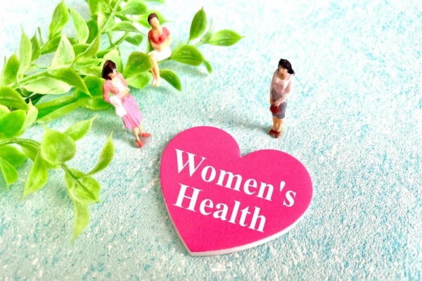 ピンク色のハート型のプレートに「Women's Health」の文字が書かれており、3人の女性のミニチュアが置かれた画像です。背景には緑の葉が飾られています。