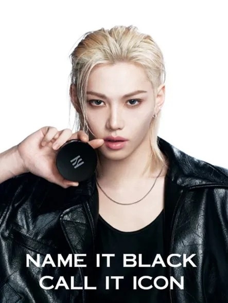 金髪の男性が黒い化粧品を持ち、黒いジャケットを着てポーズをとっている画像です。男性はカメラ目線で、テキストが画像の下部にあります。