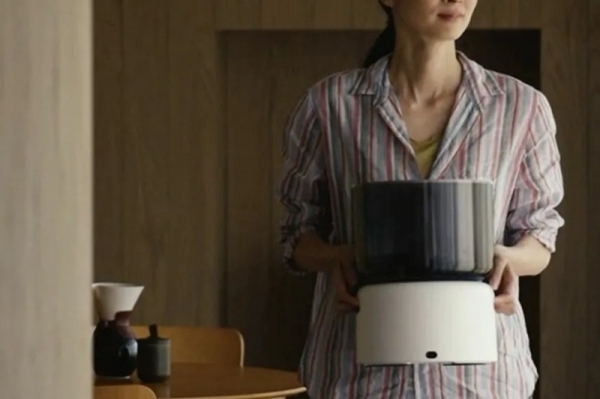 女性が白い家電のようなものを持っています。背景には、コーヒーを淹れる器具や木製の壁があります。